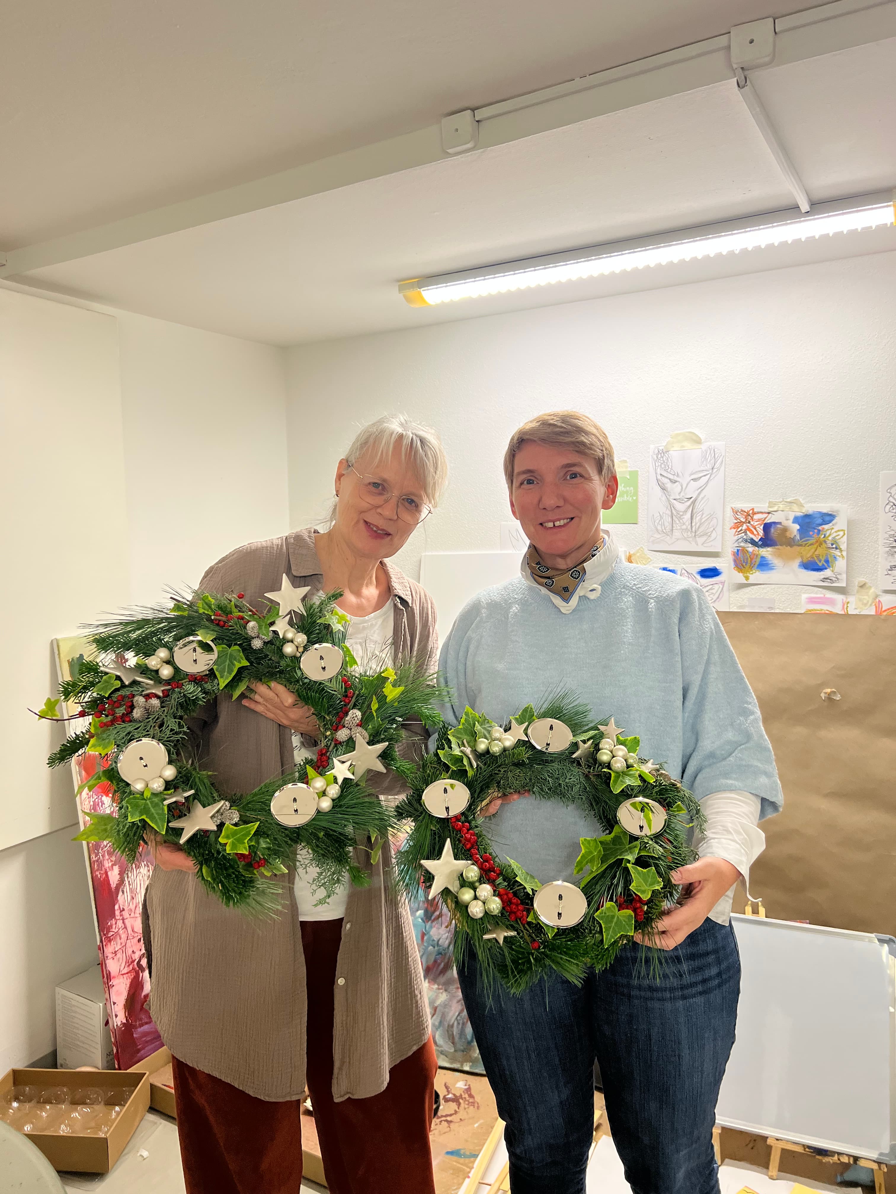 Zwei lächelnde Frauen halten ihre selbstgemachten Adventskränze in die Kamera — gemeinsame Feier im Atelier
