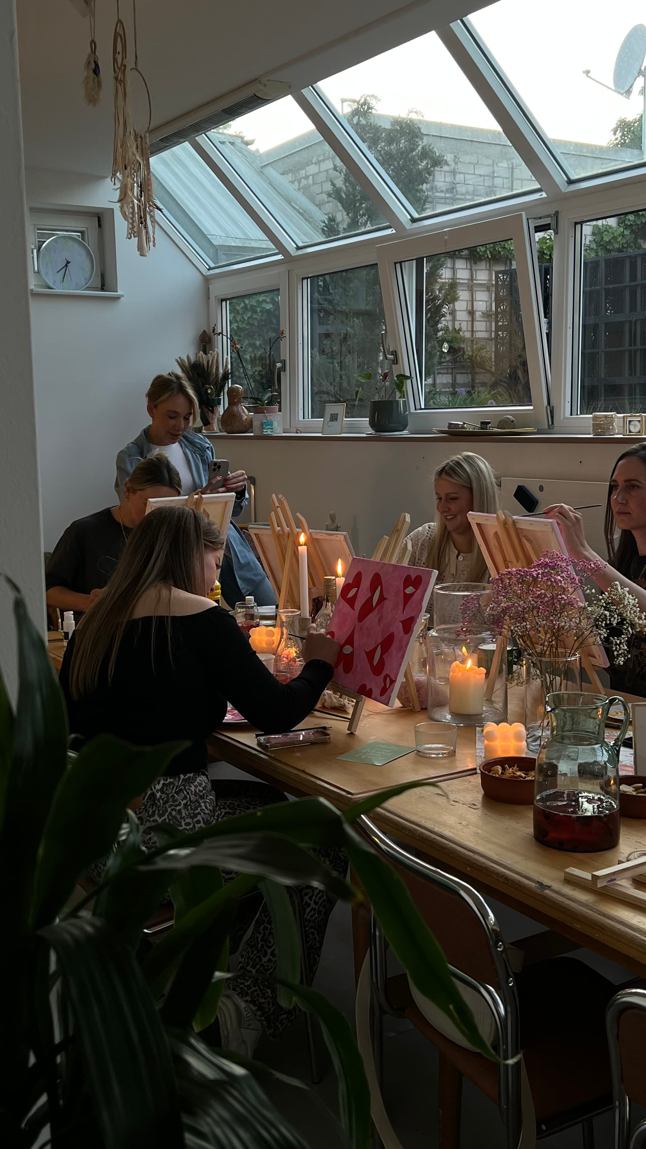 Frauen malen gemeinsam am langen Tisch unter dem Oberlicht des Wintergartens — Kerzen, getrocknete Blumen, warme Abendstimmung