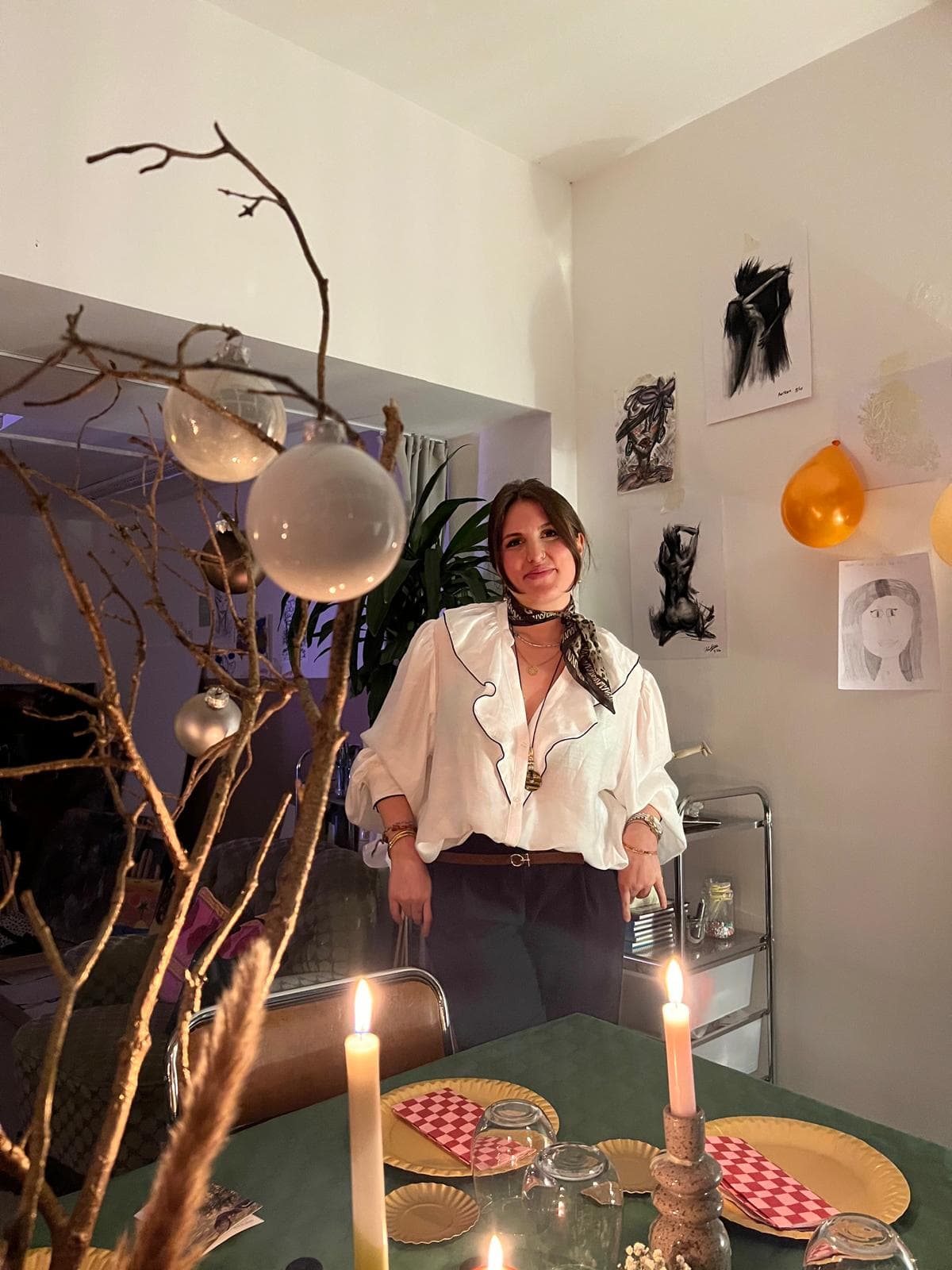 Özge Köle steht im HeimART Studio in einer cremefarbenen Bluse mit dunklem Halstuch — vor Kerzen auf dem Tisch, Zeichnungen an der Wand und einem Zweig mit gläsernen Christbaumkugeln
