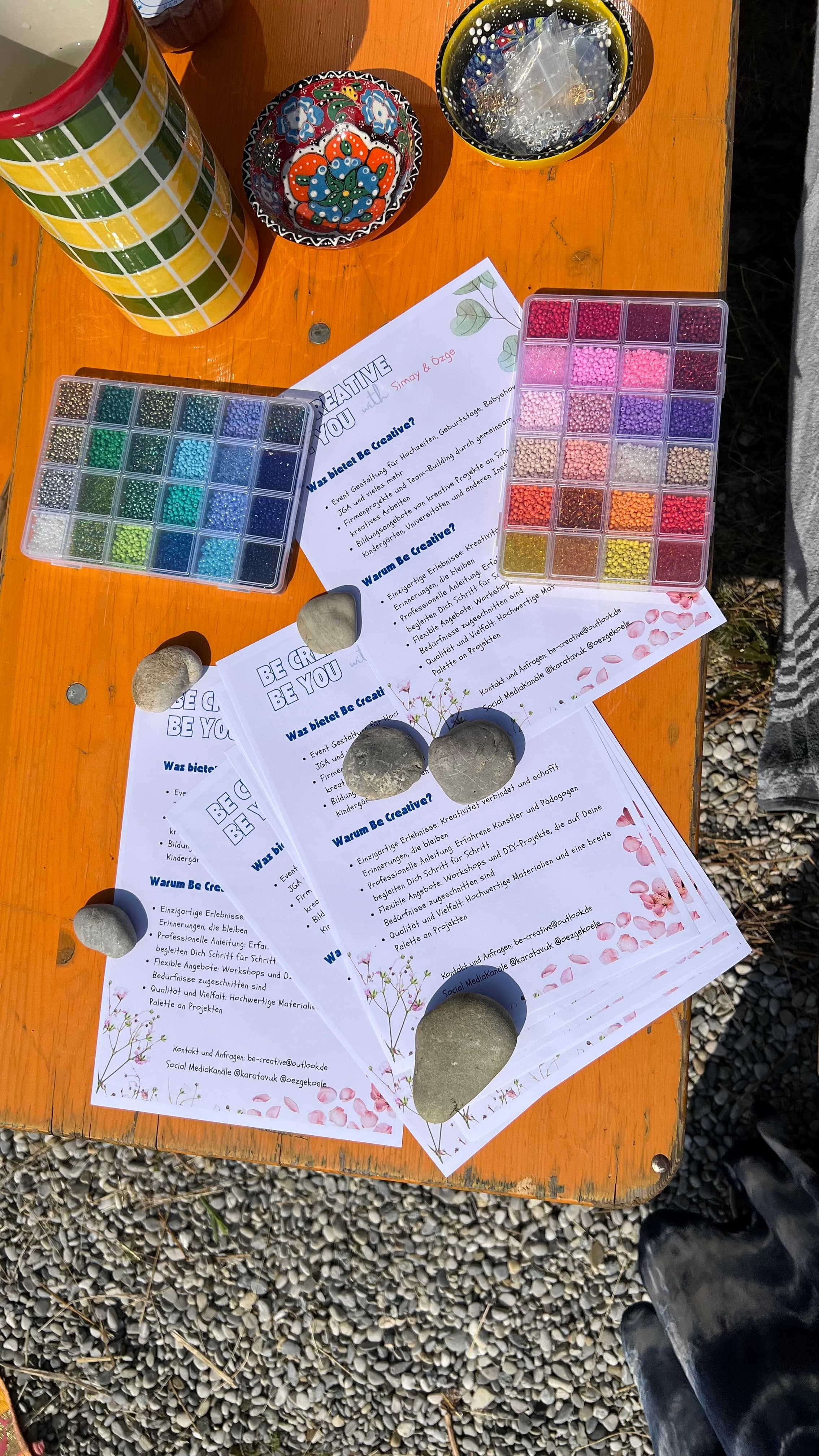 Aufsicht auf Outdoor-Tisch mit zwei Perlentabletts, Affirmations-Karten und bemalten Steinen — orangefarbenes Holz