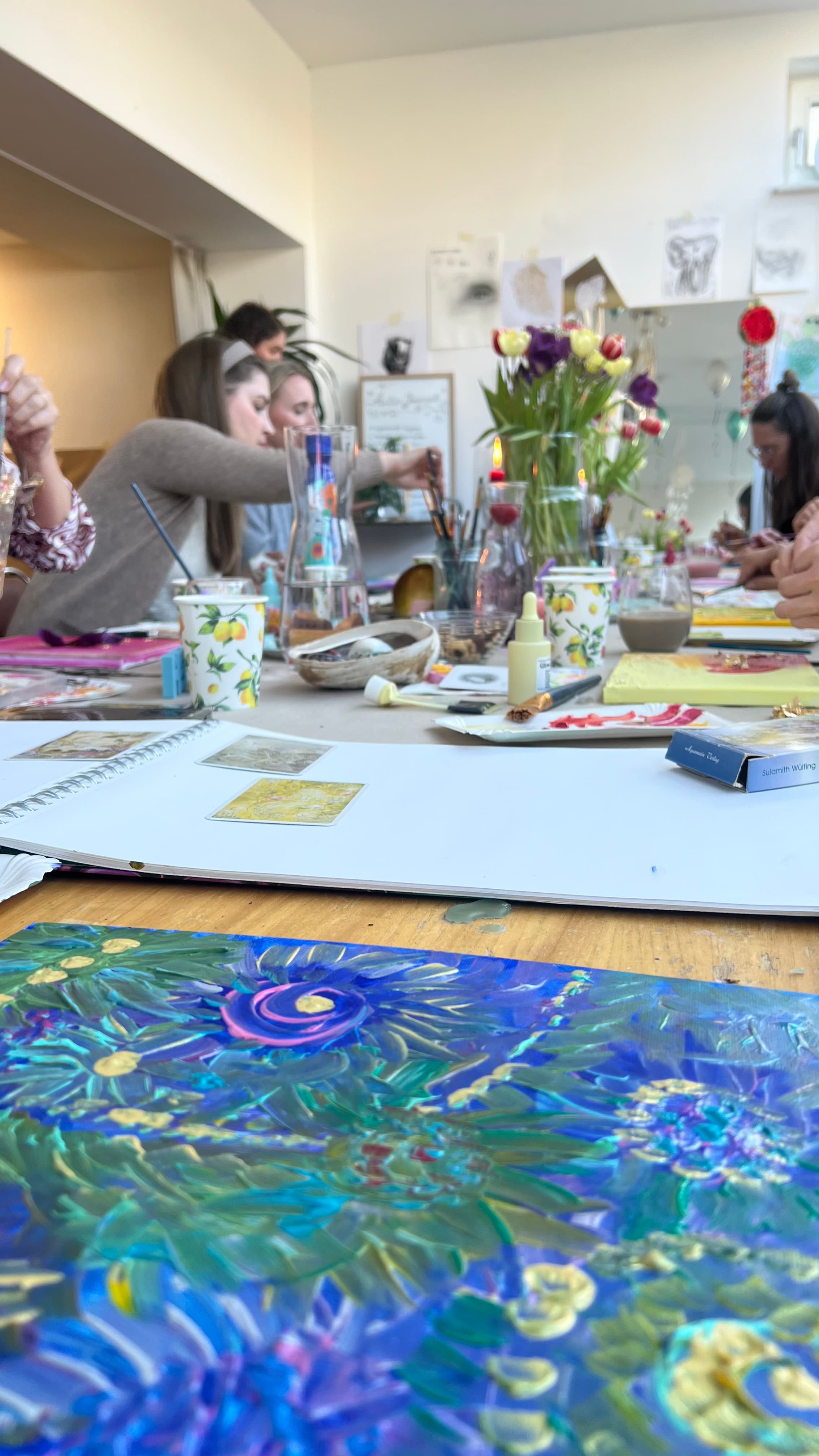 Lebendiges abstraktes Gemälde in Blau und Gelb im Vordergrund, dahinter eine Gruppe Frauen beim Malen mit Tulpen und Skizzenbüchern