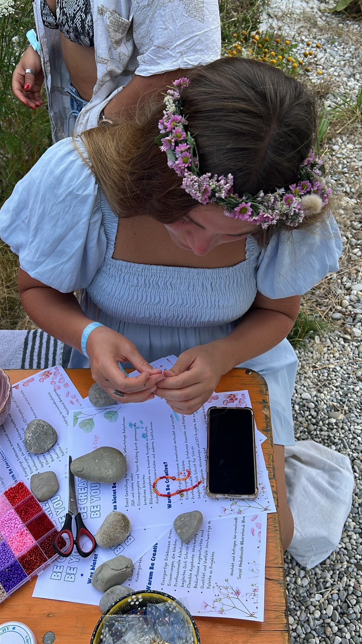 Mädchen mit Blumenkranz fädelt konzentriert Perlen auf eine Schnur an einem Outdoor-Tisch