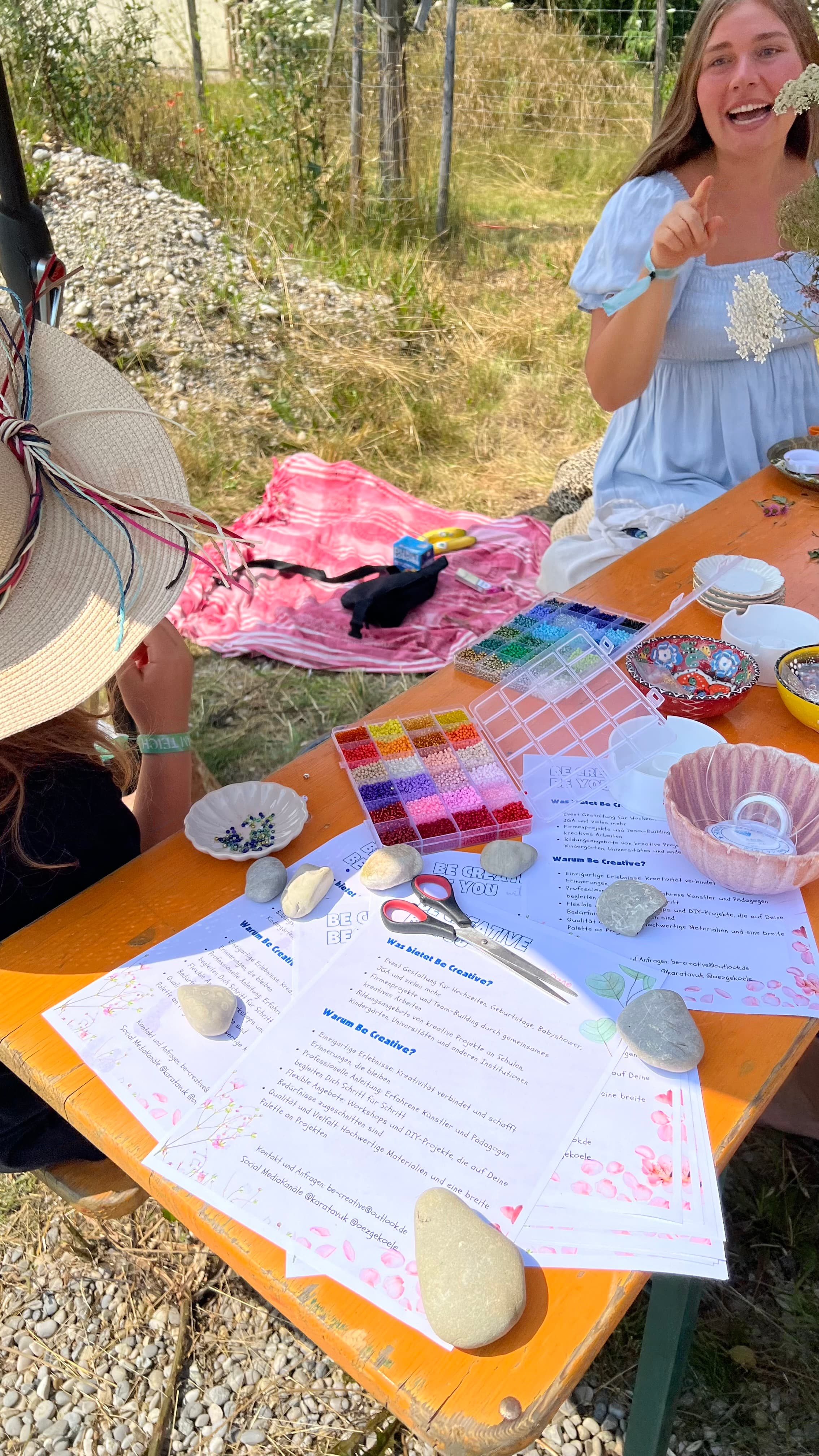 Junge Frau im hellblauen Kleid lacht herzlich an einem Outdoor-Tisch mit Anleitungsblättern, Hut und bemalten Steinen