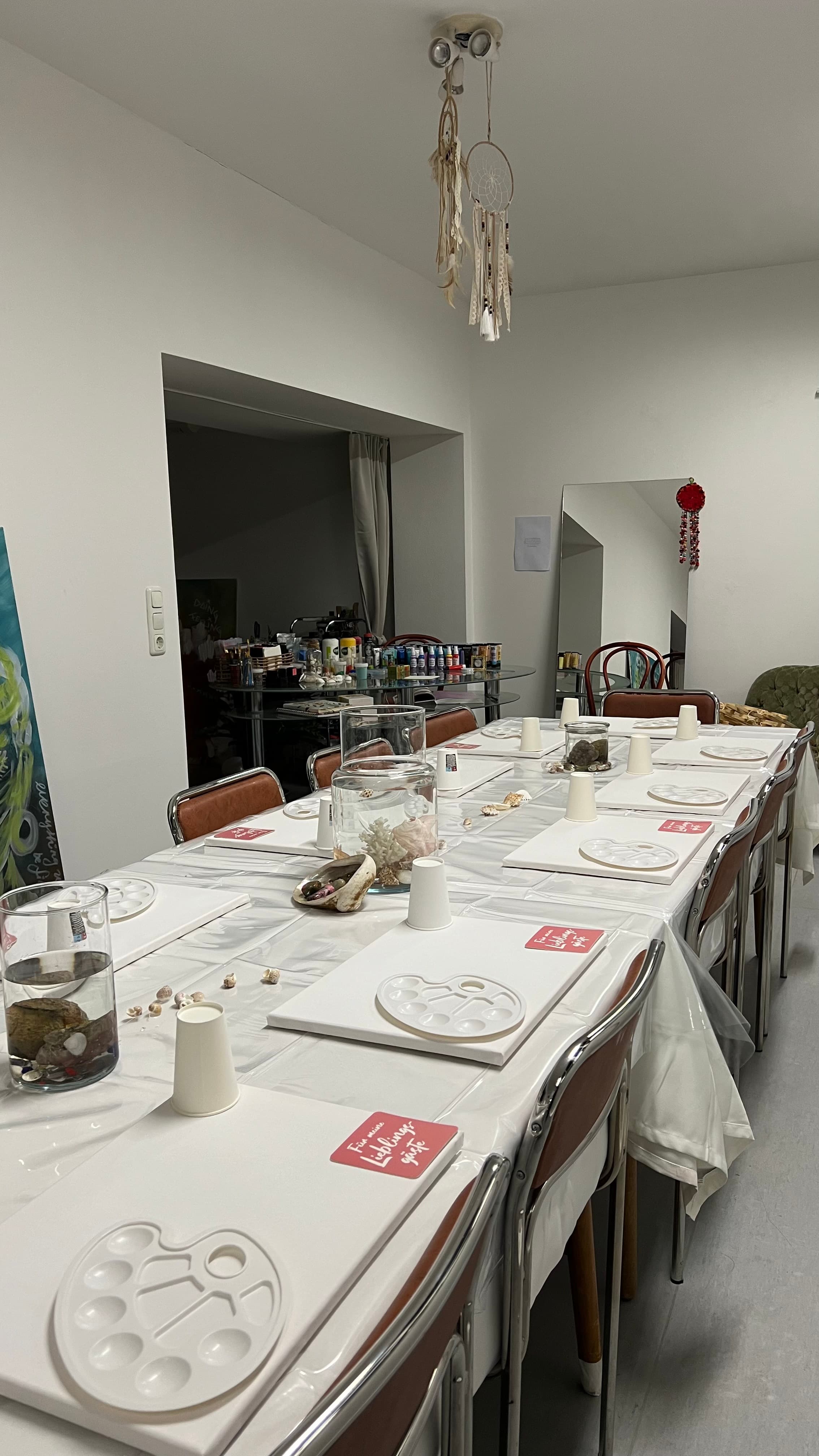 Leerer langer Tisch mit weißer Tischdecke, Paletten und Trinkgläsern unter dem Traumfänger im Wintergarten — bereit für den Abend