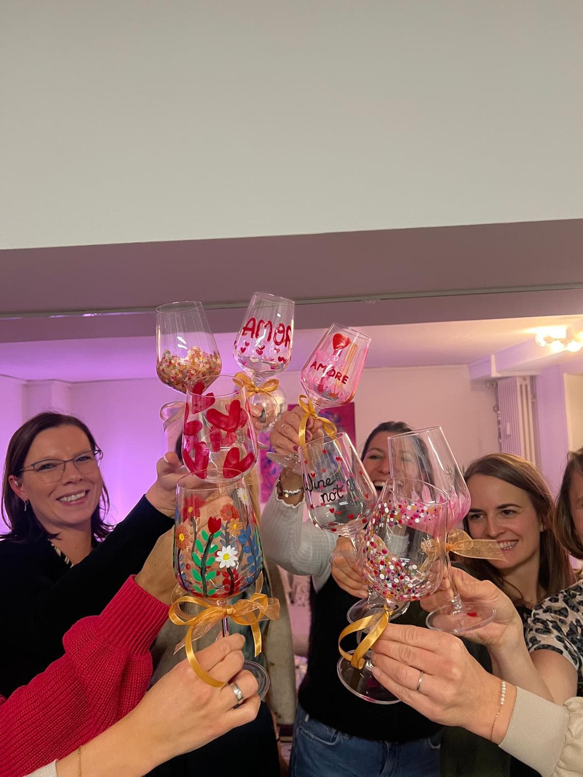 Gruppe Frauen stößt mit selbst bemalten Weingläsern an — Schriftzüge 'Amore' und 'Wine not', Herzen und Blumen, rosa Wandlicht im Hintergrund